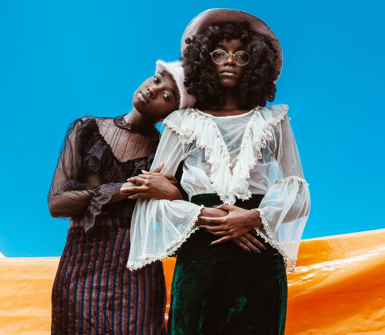 Photo of two Black women dressed in long frilly dresses and red stockings. They are leaning on each other. There is a blue, orange, and white background.