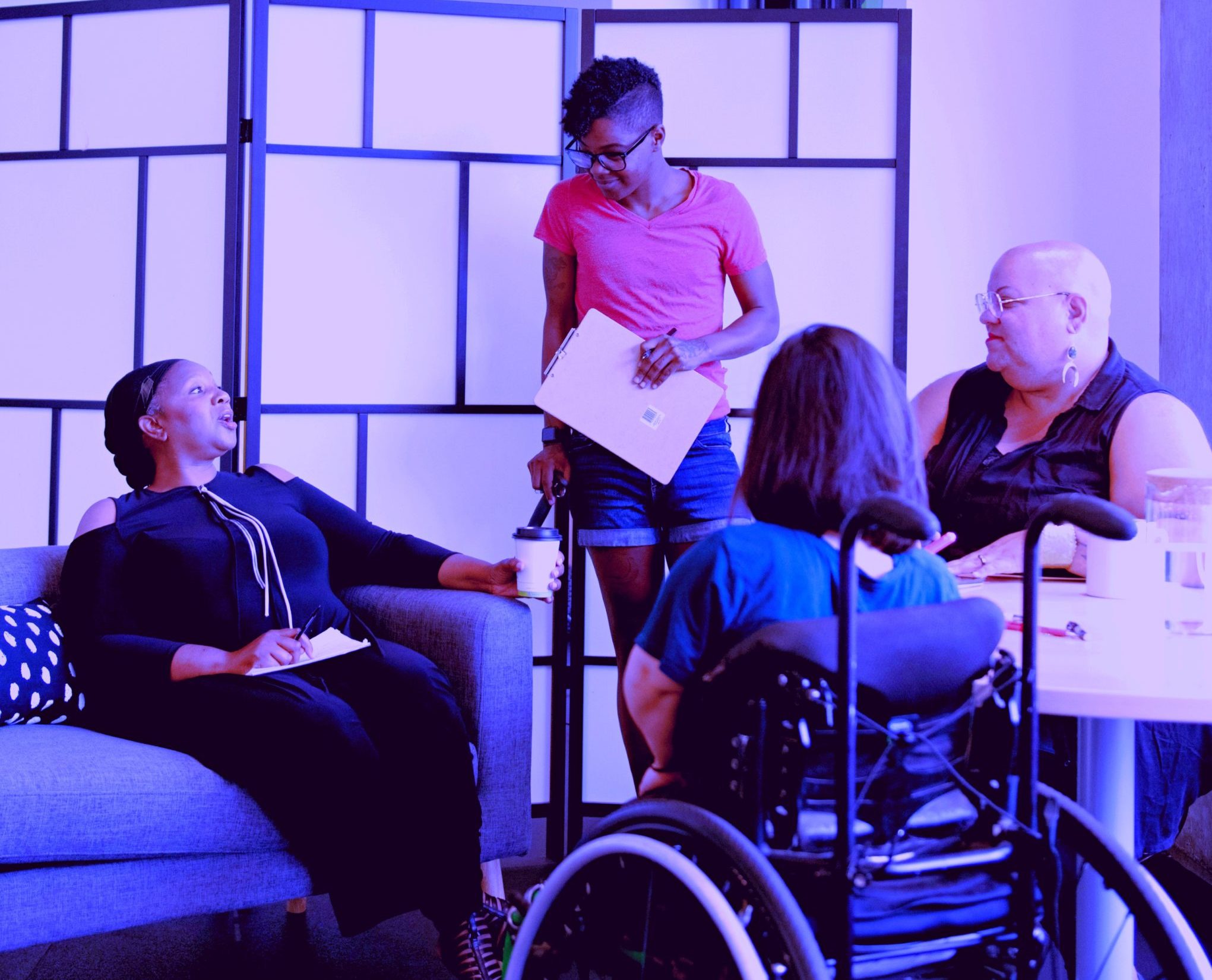 Four disabled people of color gather around a table during a meeting. A Black woman sitting on a couch gestures and speaks while the three others (a South Asian person sitting in a wheelchair, a Black non-binary person sitting in a chair, and a Black non-binary person standing with a clipboard and cane) face her and listen.