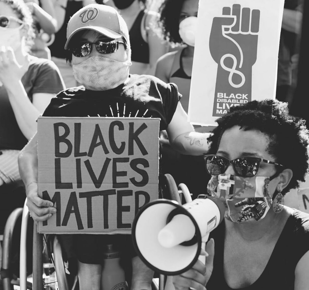 A black and white photo of Keri Gray and Mia Ives Rublee at a protest. Keri is speaking on a bullhorn and Mia is holding signs that say "Black Lives Matter."