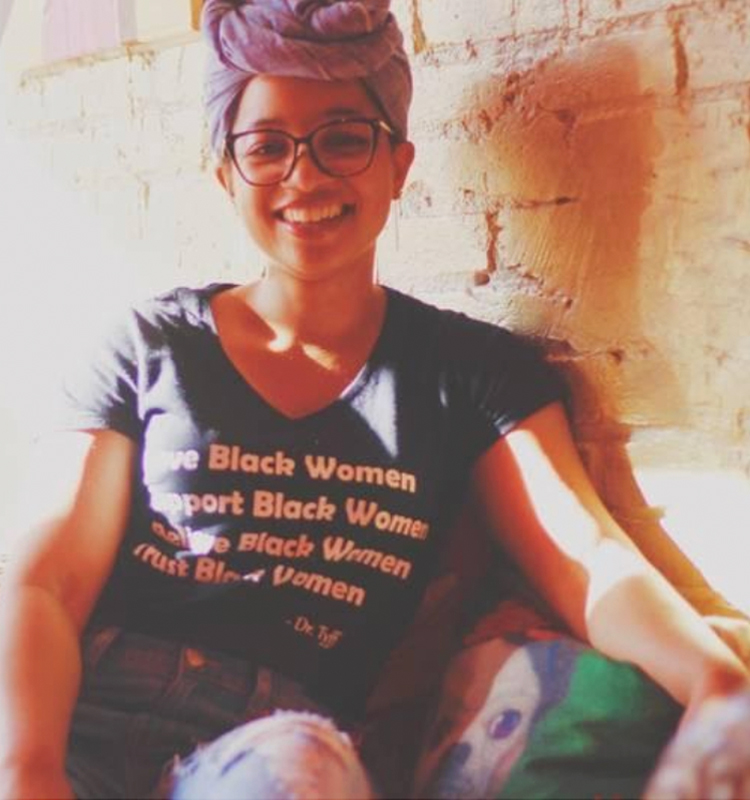 Keri Gray sitting on a couch with a brick wall behind her. She is wearing a black shirt that says Black women down the front, jeans, and a blue jean headwrap.