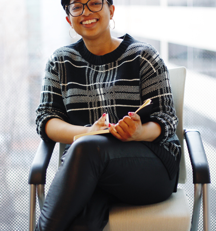 Keri Gray sitting in an office with a notepad and pen in her hand. She is wearing black leather pants and a black and white sweater.