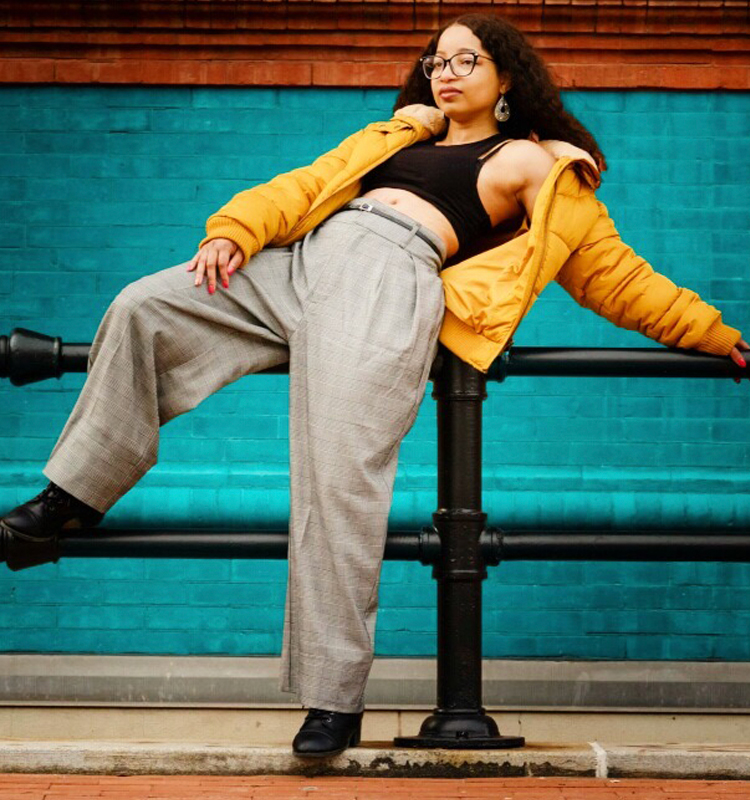 Photo of Keri wearing gray striped pants, a black cut off shirt, and a mustard coat. She is leaning against a pole with a red and blue brick wall in the background.