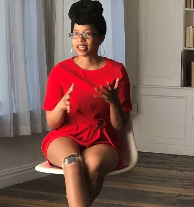 Photo of Keri Gray and she appears to be talking. She is sitting in a white chair, wearing a red romper, and a black headwrap.