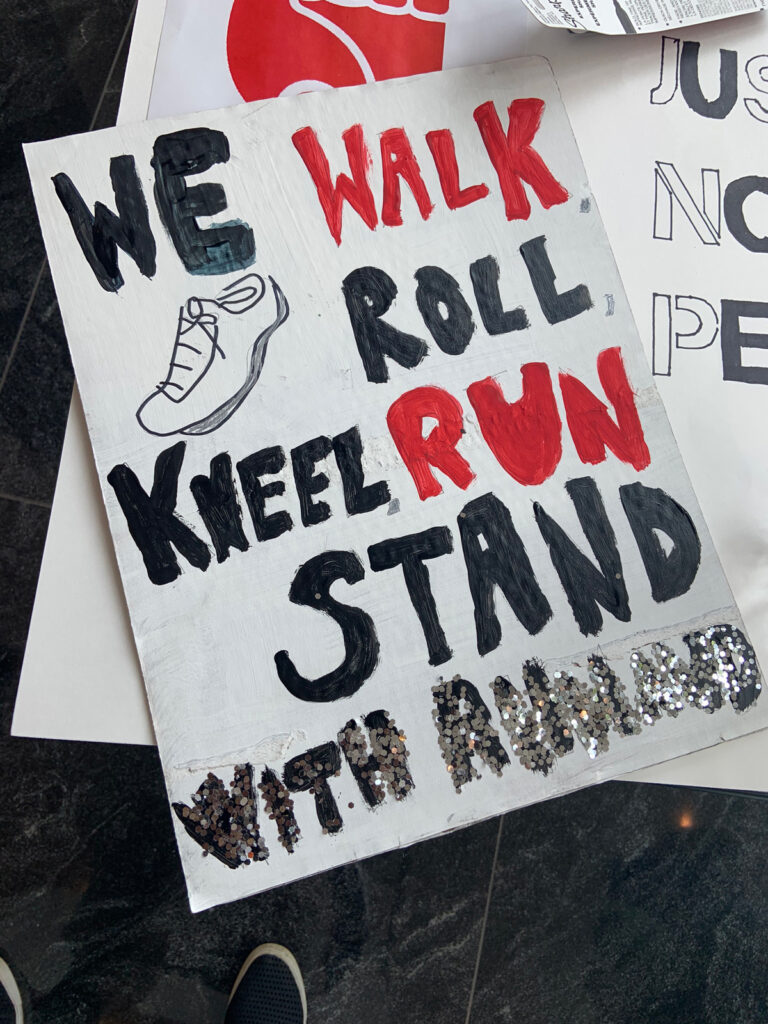 A photo of a poster that says "We walk roll kneel run stand with Ahmaud" in red, black, and silver paint.