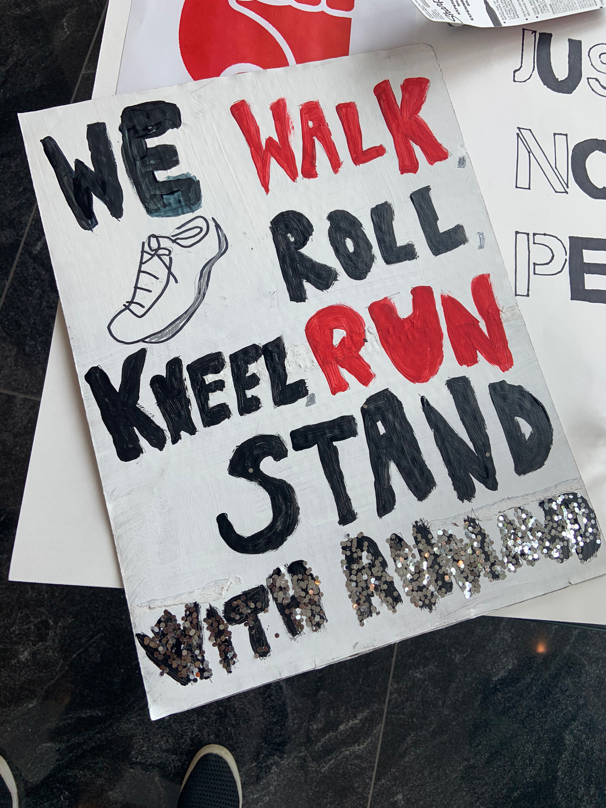 Photo of a poster that says "We walk roll kneel run stand with Ahmaud in black, red, and silver paint.