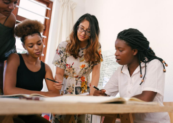 A small group of young women at a meeting. They appear to be focused on a project. On the left are two dark-skinned ladies in black tops. In the middle, stands a medium-skinned woman wearing glasses in a floral dress. On the right is a dark-skinned woman with black dreads in a white top.
