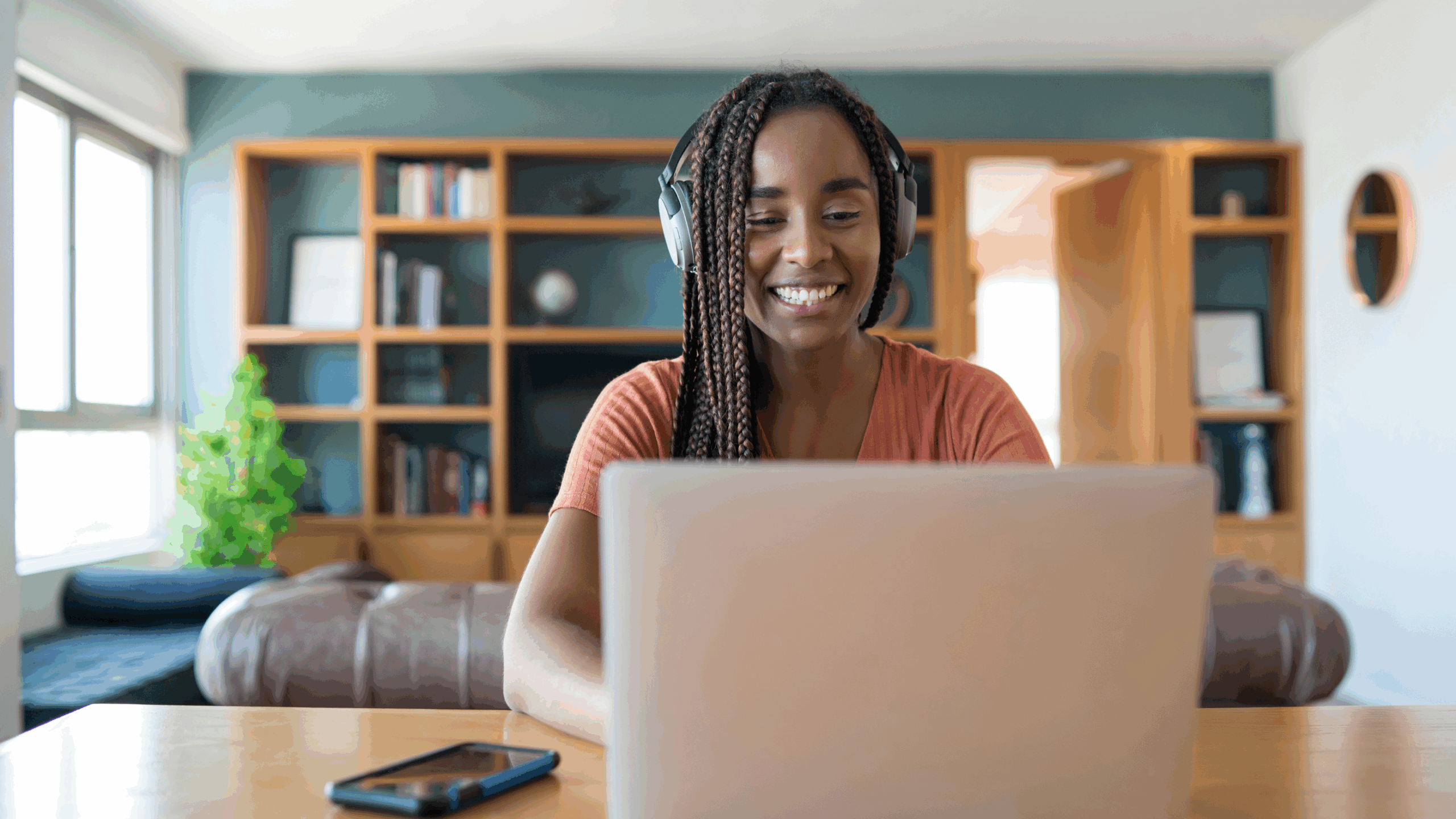 At home, a young woman with dark skin and long, brown locs is working on her laptop while wearing headphones. She's smiling.