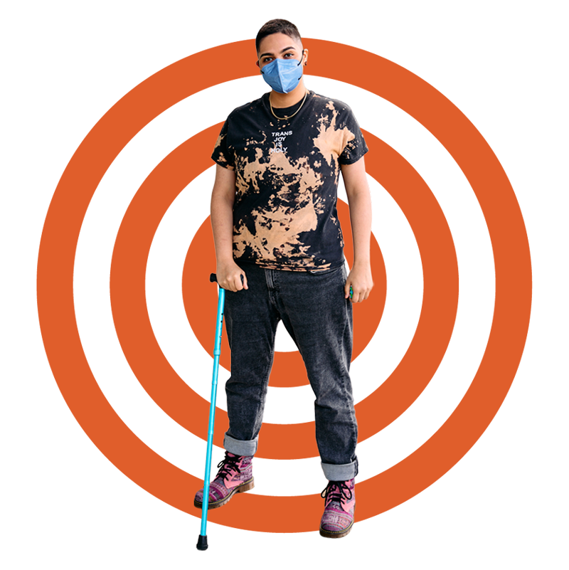 A person of color using a cane stands confidently against a bold orange concentric circle graphic on a black background, wearing a mask and a shirt that reads "Trans Joy is Holy"