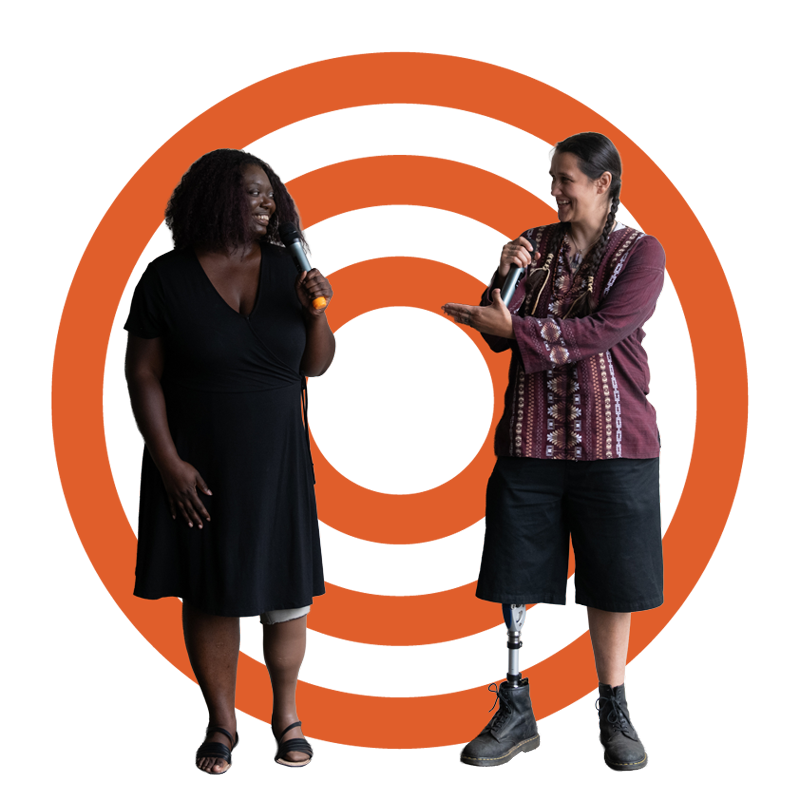 An Indigenous Two-Spirit person and a Black woman face each other and gesture while smiling and speaking into microphones. They are both amputees donning prosthetic legs.