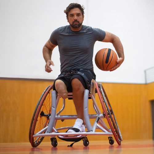 A person using a sports wheelchair dribbles a basketball on an indoor court, leaning forward in an athletic stance
