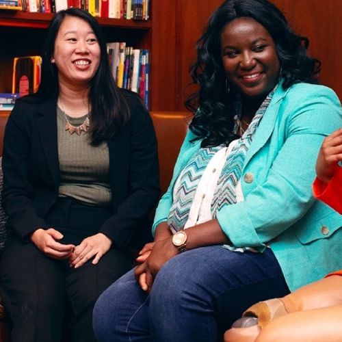 Two people of color — an Asian person and a Black person — sit together smiling in a room lined with bookshelves