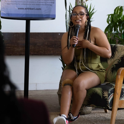 A Black person with long braids and a prosthetic leg sits in a chair holding a microphone and speaking at a community event, with a banner and plants visible in the background