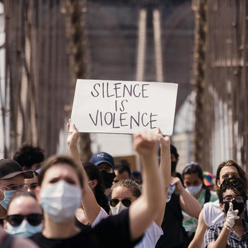 A crowd of masked protesters marches across a bridge, with one person holding a sign that reads "Silence is Violence"