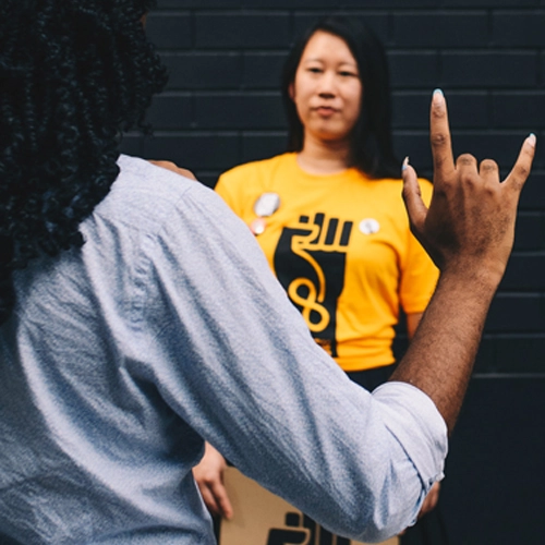 A person of color signs in the foreground while another person in a NAMED Advocates shirt stands in the background against a brick wall